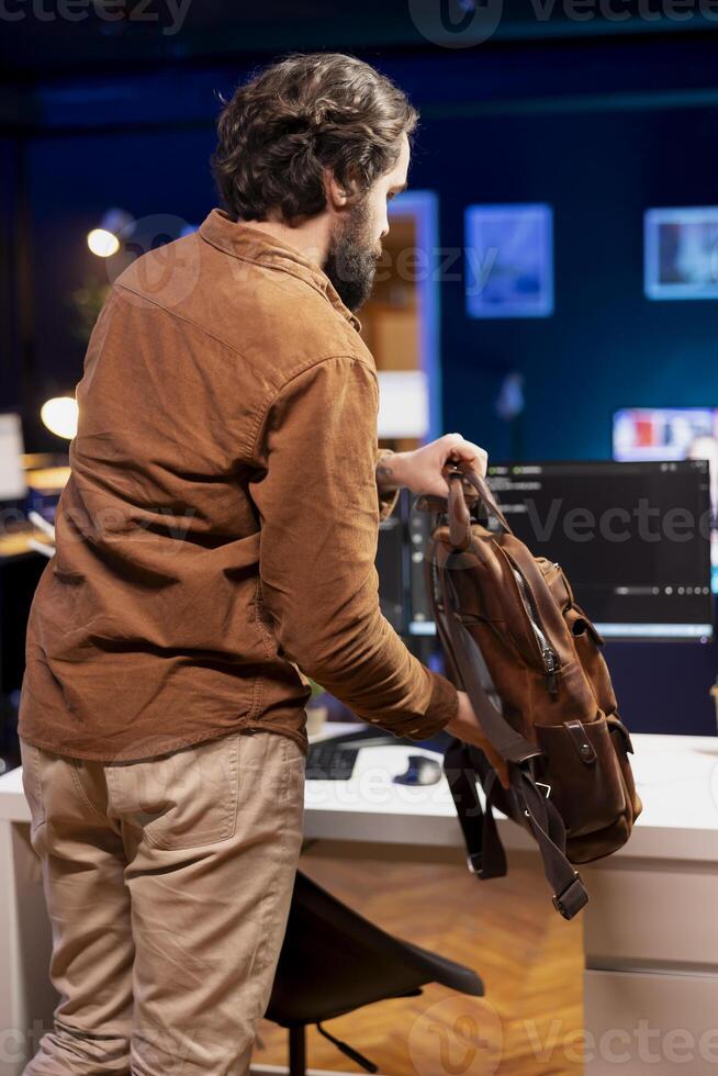 Programmer packing backpack, leaving office after implementing security measures, protecting company systems and data. IT worker ending work shift after finishing tasks and debugging code photo