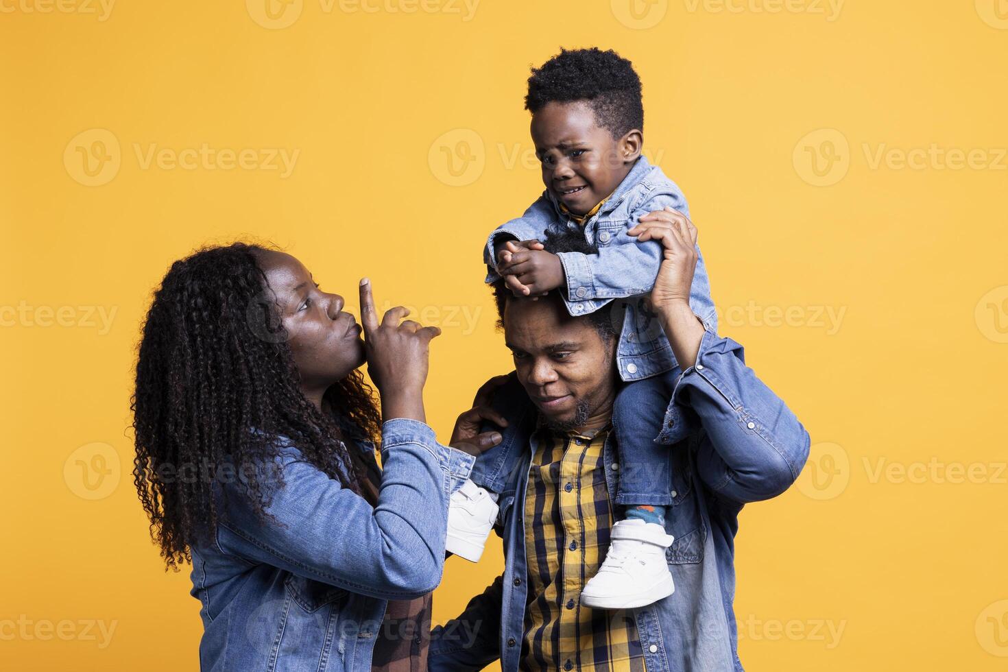 African american parents calming down their infant son while he cries, his father is carrying him on his shoulders. Young mother showing hush sign to his toddler, trying to make him smile. photo