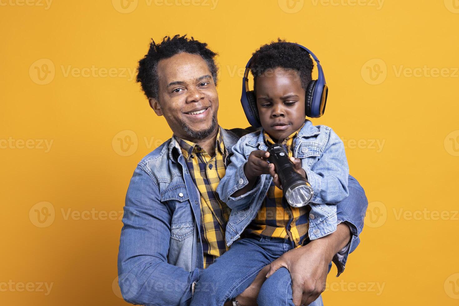African american dad and his child playing with a flashlight on camera, sweet boy wearing headset while his father is carrying him around the studio. Adorable kid discovering a lamplight. photo