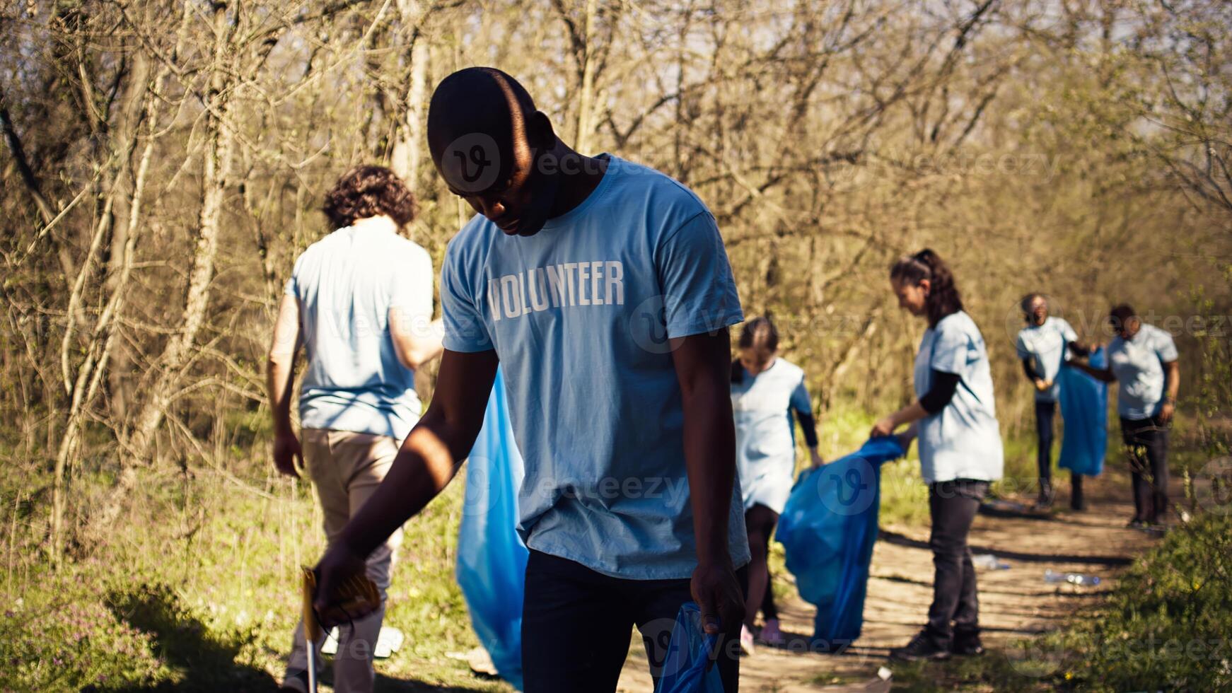 Eco friendly activist grabbing garbage and plastic waste from a forest area, cleaning up the woods and collecting rubbish in a trash bag. Man volunteer dealing with waste management. Camera B. photo