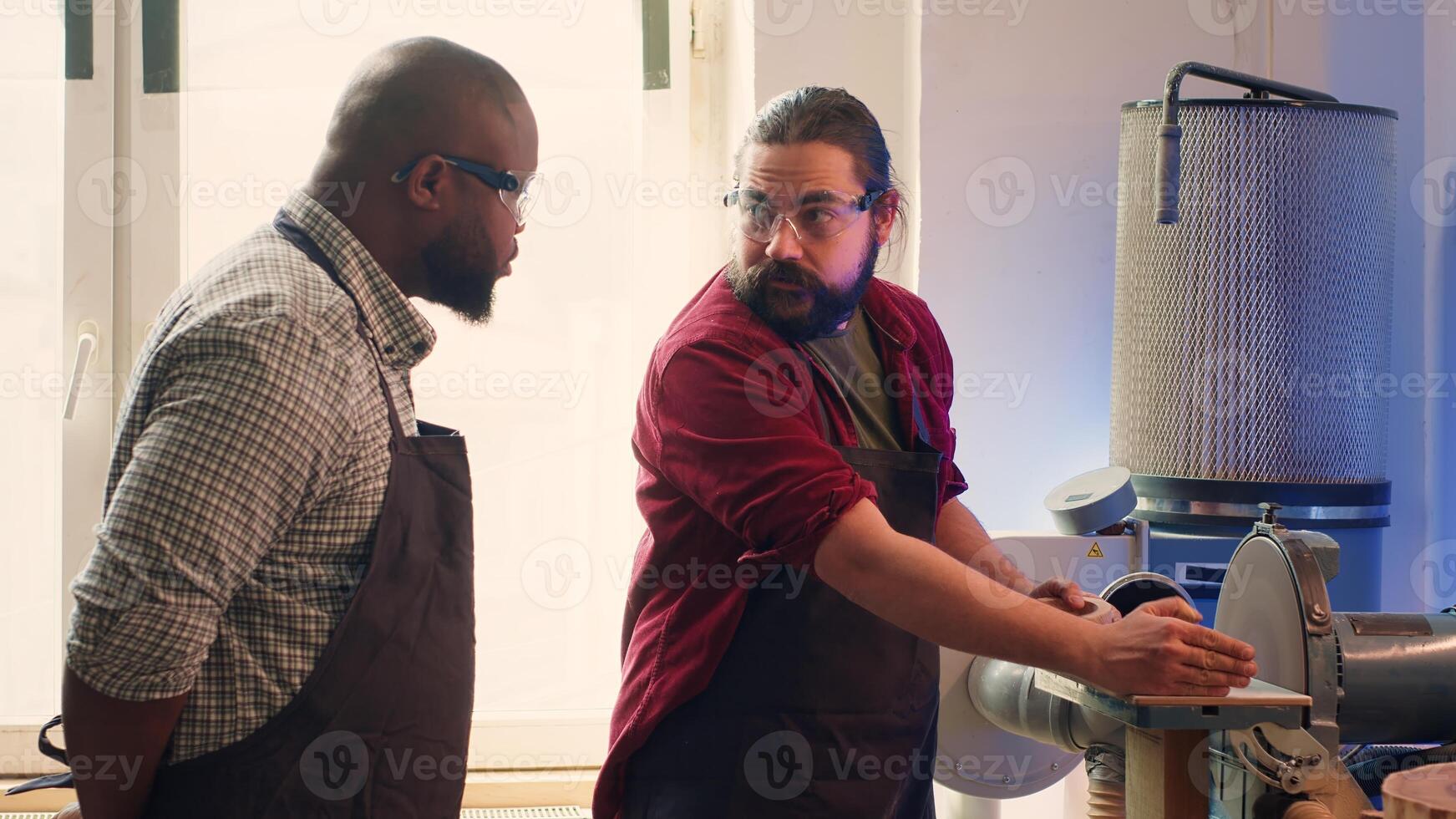 Manufacturer teaching new employee how to shape wooden bowl on disc sander in assembling shop. Apprentice learning from woodworking specialist how to use grinding machine rotating disc, camera A photo