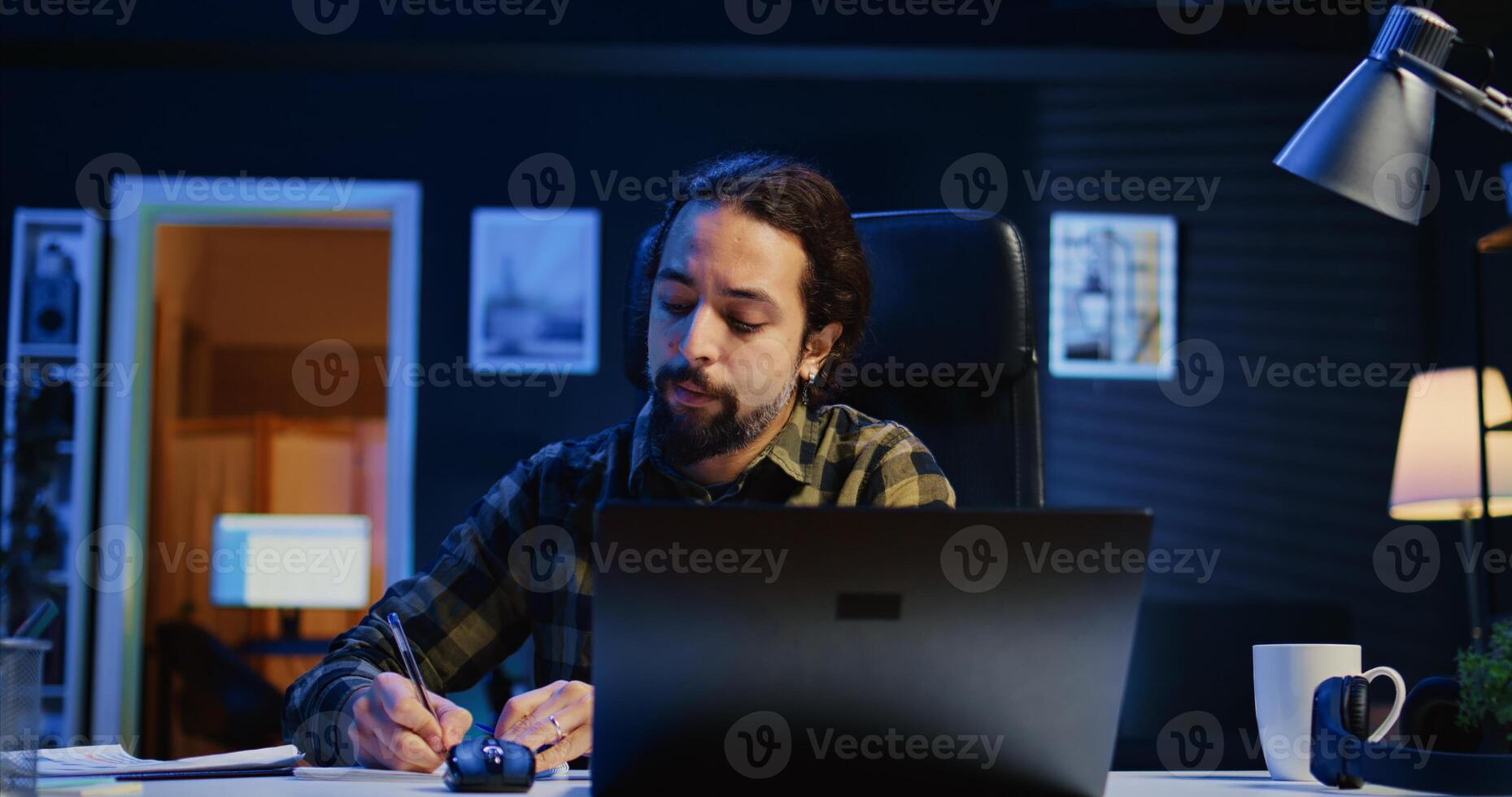 Entrepreneur working at home office desk on laptop, browsing the internet and writing in notebook. Caucasian man using pen, notepad and digital device while teleworking, camera B photo