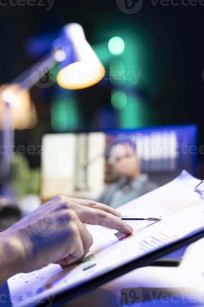 Closeup of person reviewing information on the clipboard while being part of virtual meeting on laptop. Selective focus of an individual checking charts and graphs while on call. photo