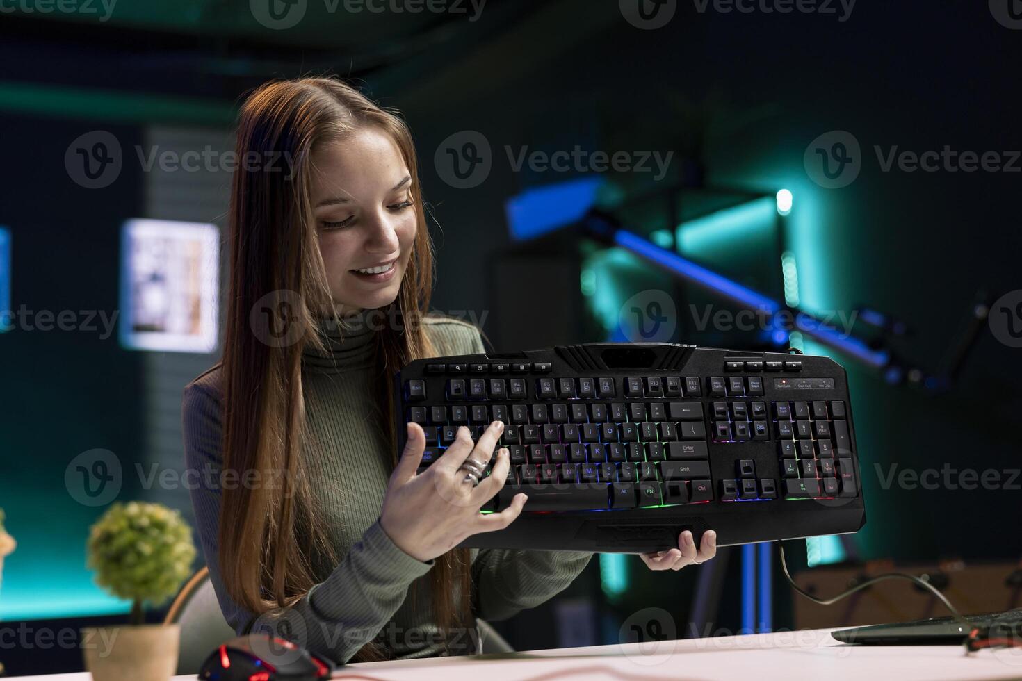 Joyous girl reviews RGB lit keyboard from sponsor for online streaming channel in apartment. Smiling media star excited to promote gaming computer peripherals brand in neon lit studio photo