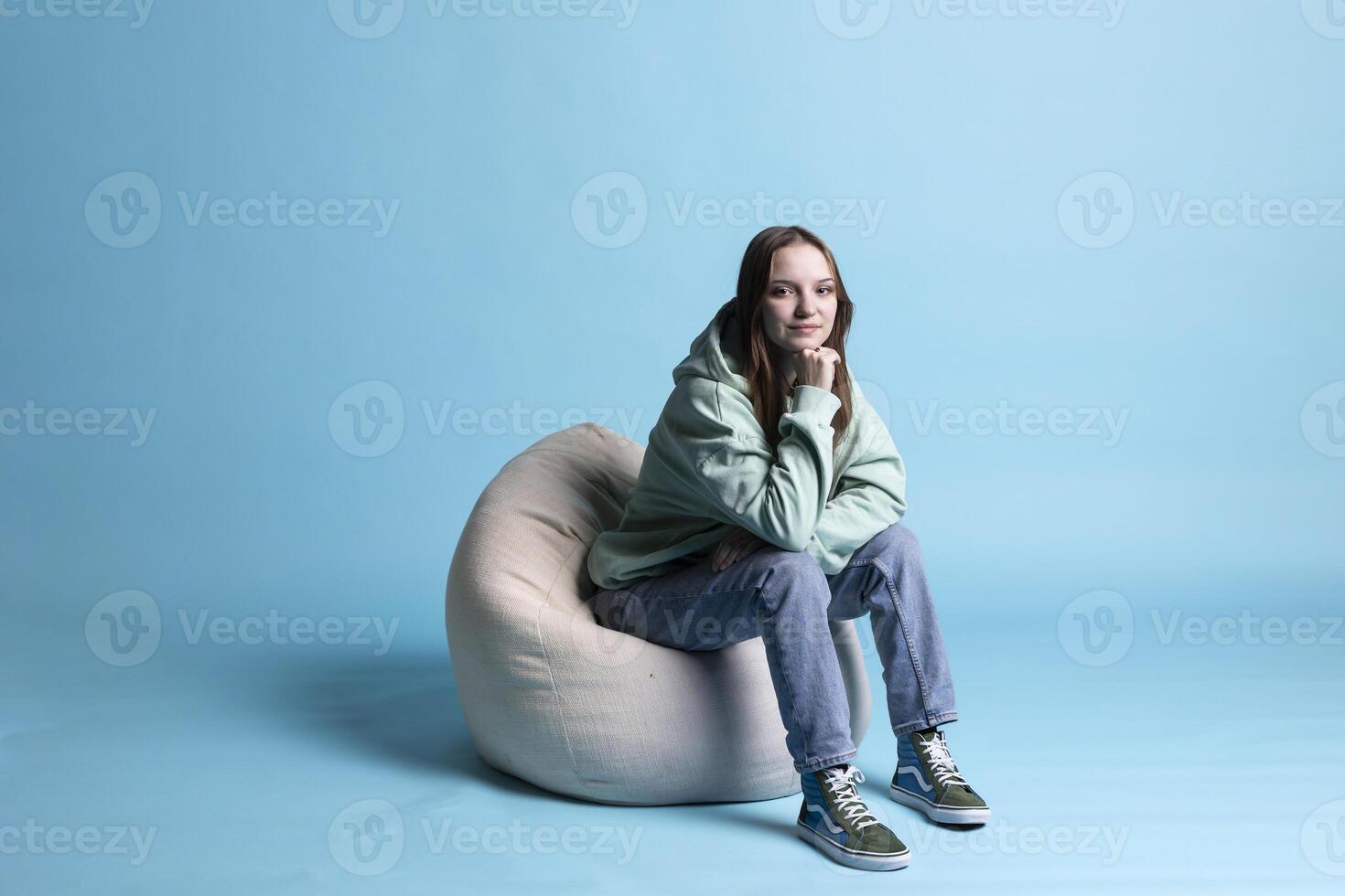 Portrait of happy girl sitting on beanbag armchair and smiling, isolated over blue studio background. Upbeat teenager seated on cozy bean bag, resting at home, having positive mood, studio backdrop photo