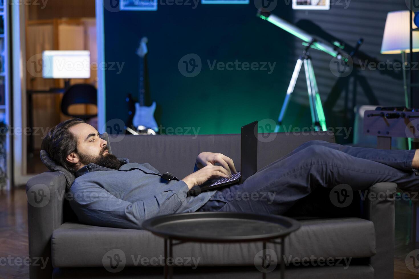 Freelancer lounged on couch working from home in apartment solving business related tasks on laptop. Remote employee laying down in neon lit living room, sending emails on notebook photo