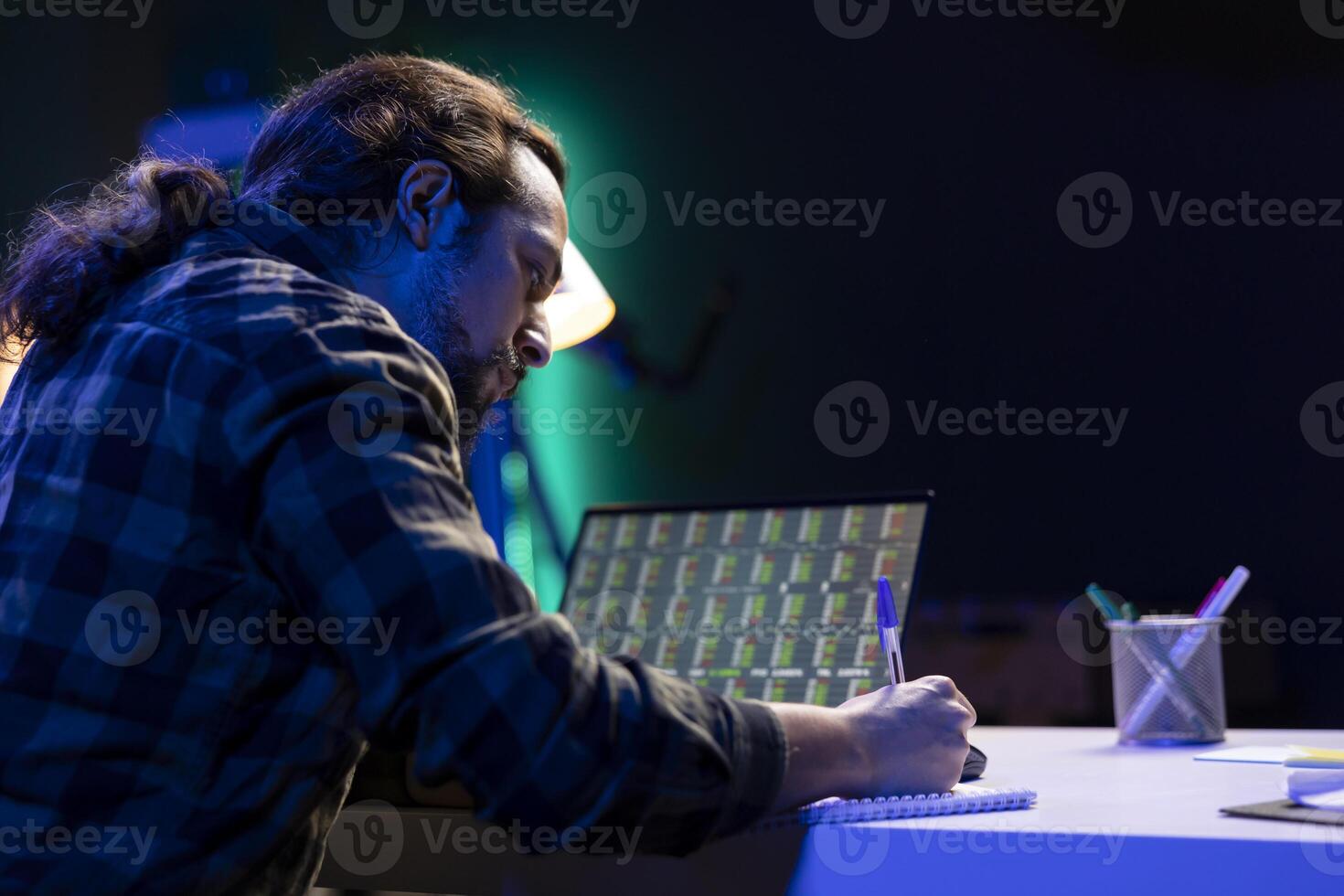 Hardworking man multitasking, looking over a forex chart on his laptop and taking notes. Male freelancer checking the stock market trend values on his device while working from home with a notebook. photo