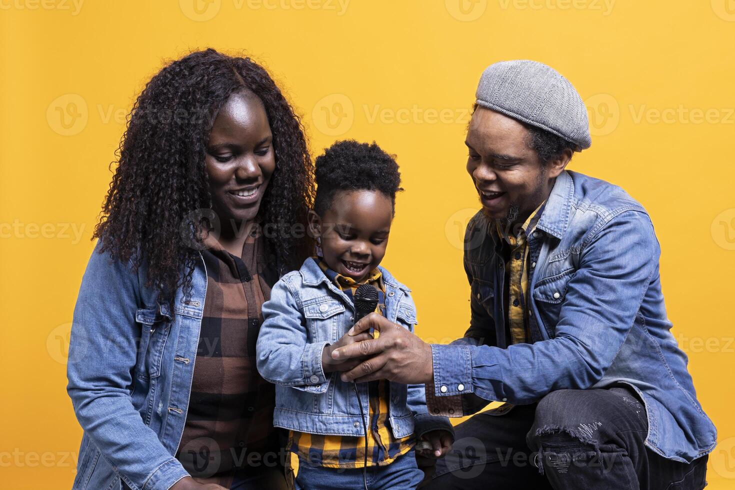 Proud parents listening to their infant son singing a song on microphone, laughing and being adorable together against yellow background. Happy african american family of three enjoy music. photo