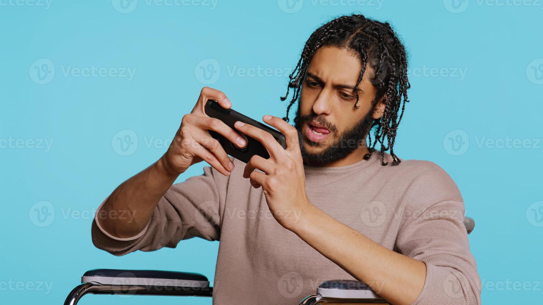 Focused man with disability plays intense videogame on mobile phone, spamming attack buttons on touchscreen. Gamer participates in gaming competition on wheelchair, studio background, camera B photo