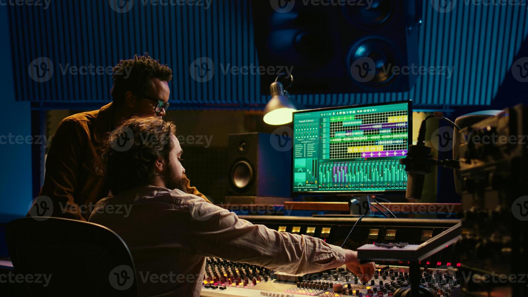 Musician and producer work on soundboard pads at professional studio station. Sound designer uses digital mixer in music production, operating on sliders and key buttons at control desk. Camera B. photo