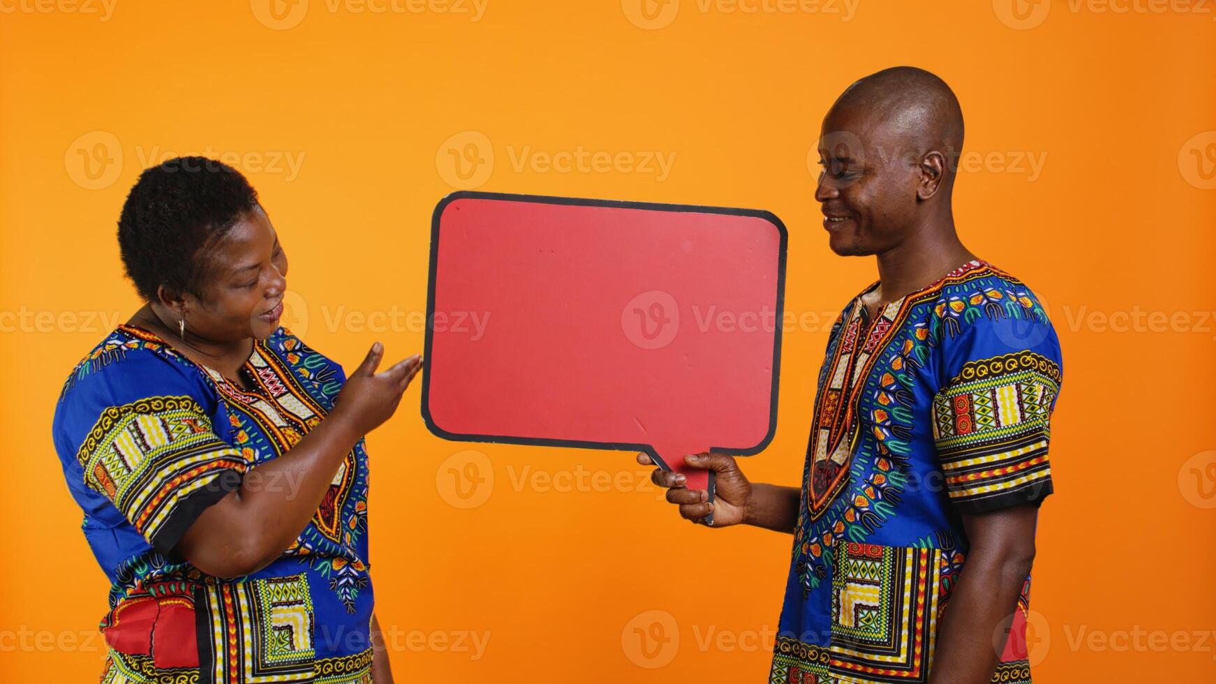 Ethnic young couple presenting speech bubble board, creating marketing advertisement and promoting red billboard empty sign in studio. African american people pointing to cardboard icon. photo