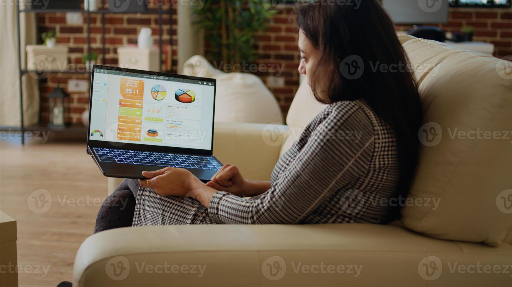 Remote employee sitting on couch in apartment, optimizing market performance charts while working from home. Woman checking online business analytics statistical data metrics, camera A panning shot photo