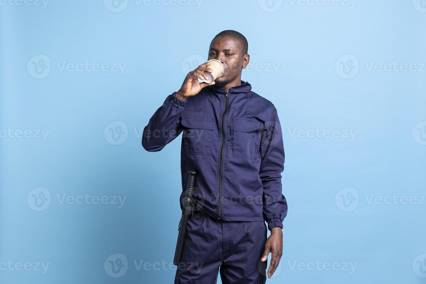 Portrait of african american security agent drinks a cup of coffee in studio, enjoying a caffeine refreshment against blue background. Confident armed employee serving a beverage. photo
