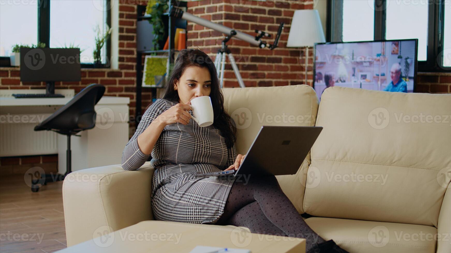 Teleworker lying on couch, drinking coffee while remotely performing tasks in personal office. Freelancer feeling stressed, enjoying hot beverage to relax while inputting data on laptop, camera A photo