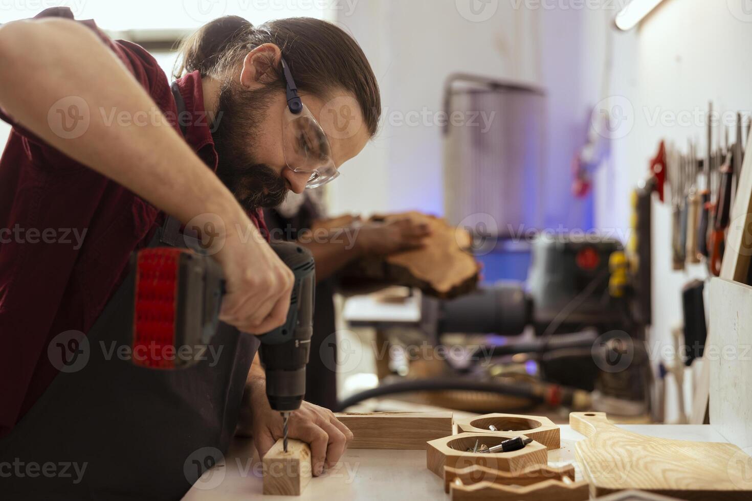 Man in carpentry shop wearing protection gear while drilling holes into wood to prevent workplace accidents. Cabinetmaker equipped with safety glasses while using power drill to avoid injury photo