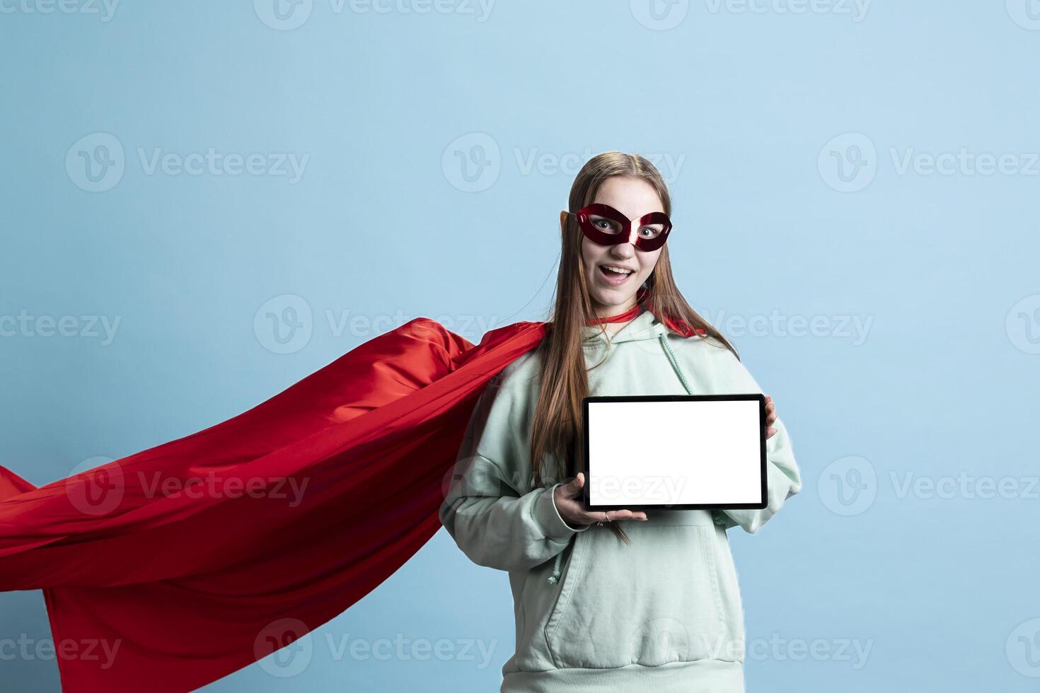 Joyful girl wearing superhero costume and mask for Halloween, using isolated screen tablet, studio background. Portrait of radiant person dressed as comic book hero holding mockup digital device photo