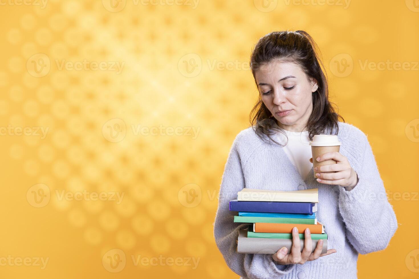 Woman holding pile of books, enjoying cup of coffee in the morning. Tired person with stack of novels in arms, drinking caffeinated beverage, isolated over studio background photo