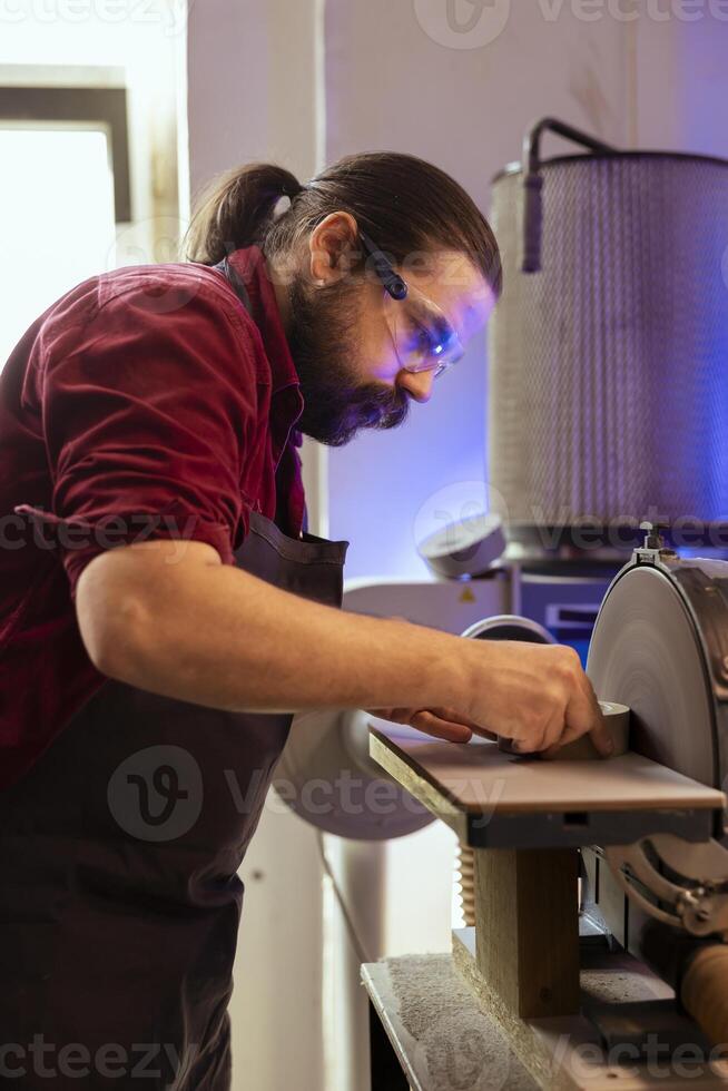 Carpenter in woodworking shop wearing protection goggles while using disc sander to avoid injury. Cabinetmaker in joinery using professional grinding machine with safety gear on to prevent accidents photo