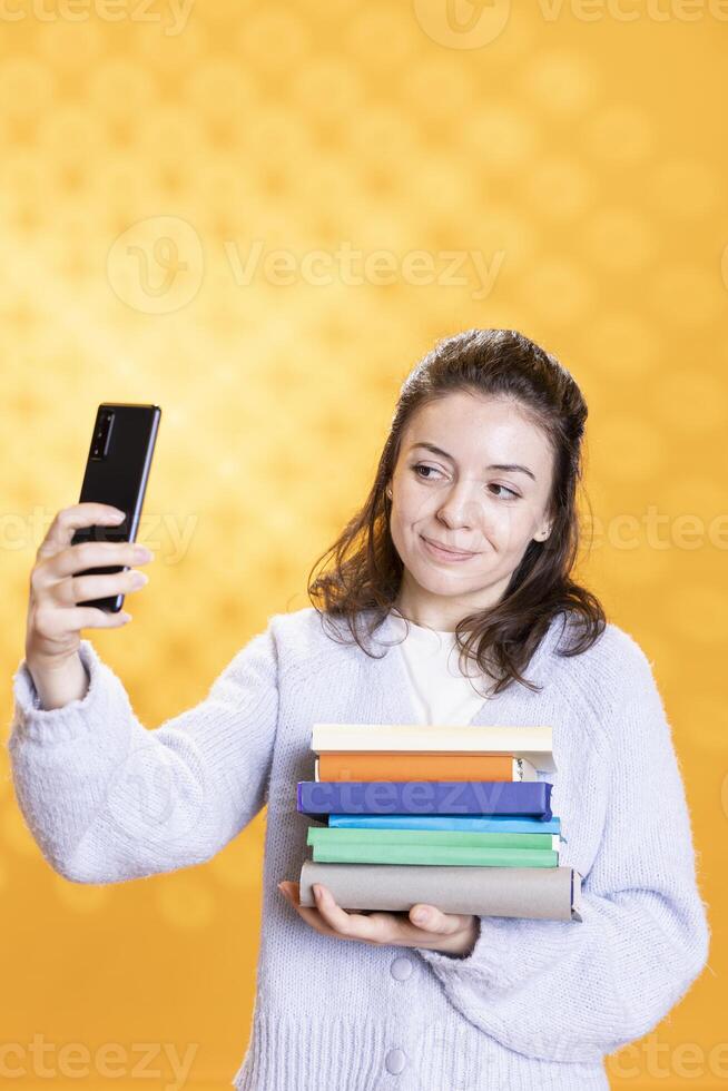 Smiling woman holding pile of books, enjoying reading hobby, taking selfies with cellphone. Cheerful lady with stack of novels in arms doing pictures with mobile phone, studio background photo