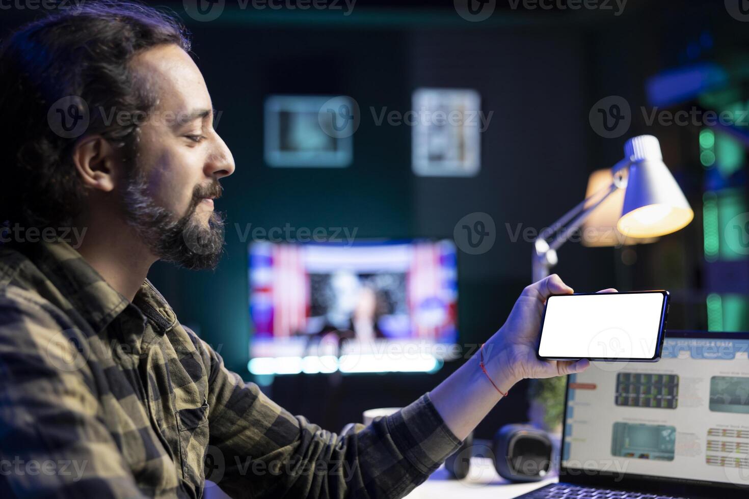Man having a smartphone showing isolated copyspace mockup template. Male freelancer working at home on a laptop while holding his mobile device displaying blank chromakey white screen. photo
