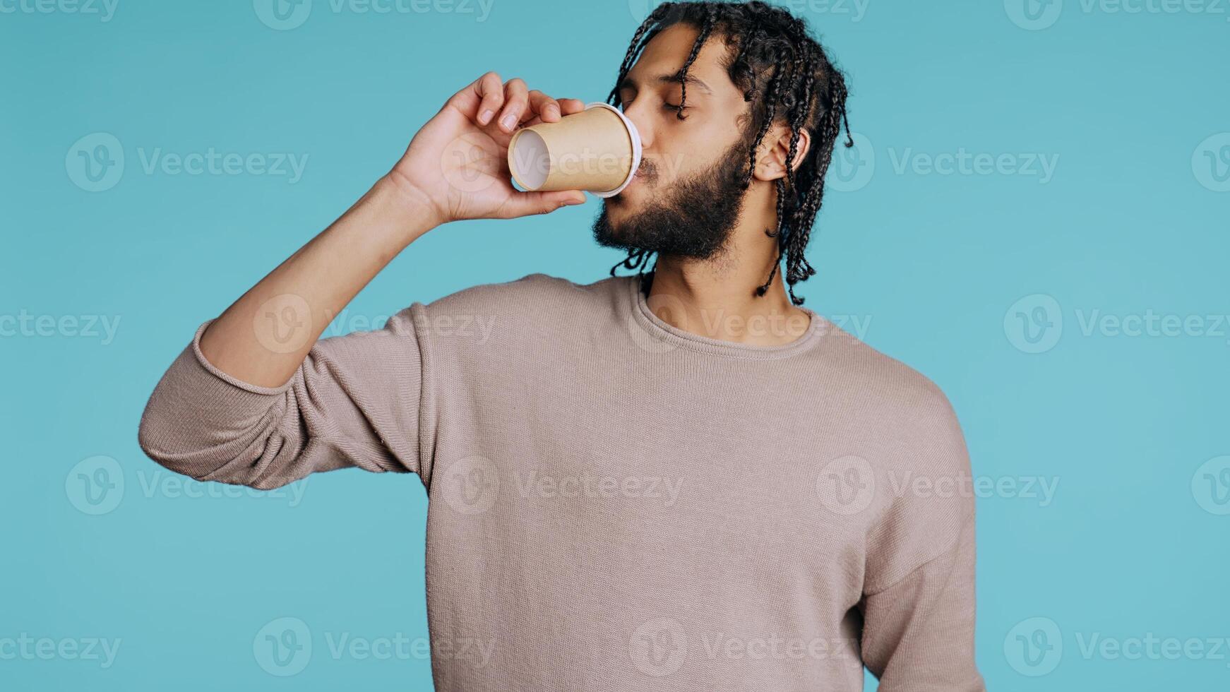 Man enjoys fresh coffee from disposable paper cup early in the morning to wake up and be energized. Person drinks hot beverage from brown recycled takeaway cappuccino cup, studio background, camera B photo