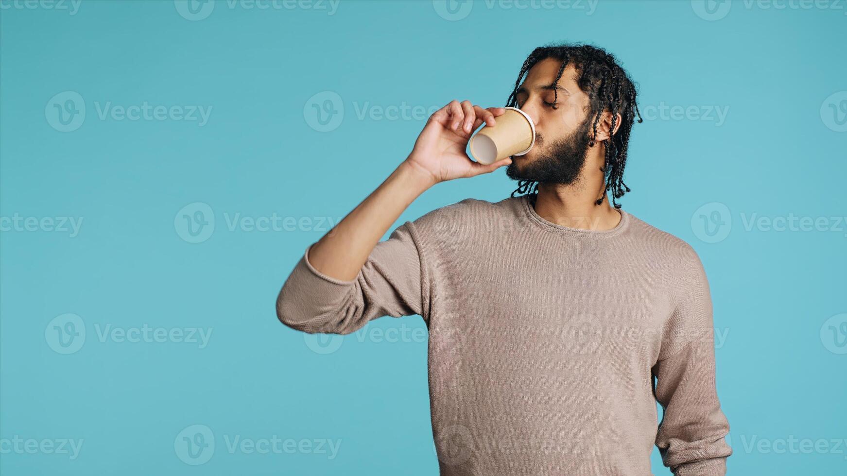 Man enjoys fresh coffee from disposable paper cup early in the morning to wake up and be energized. Person drinks hot beverage from brown recycled takeaway cappuccino cup, studio background, camera A photo