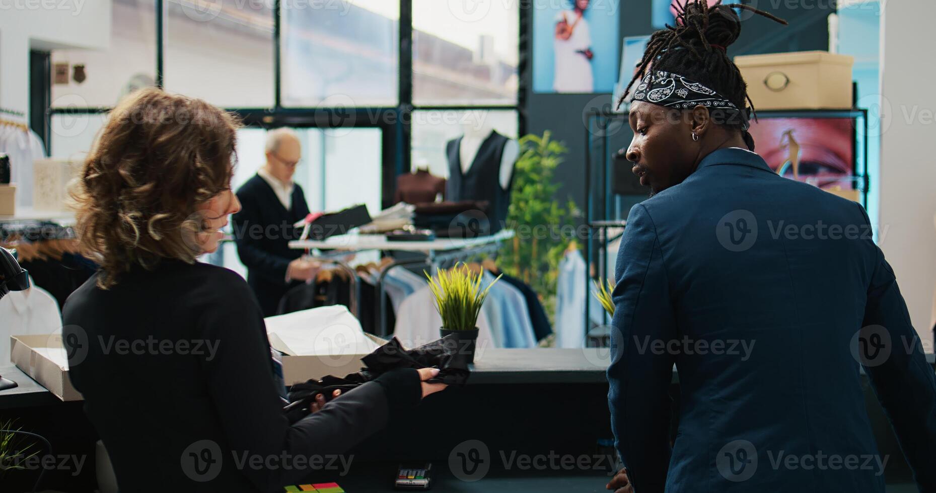 Store assistants receiving new fashion collection to put on display in showroom, looking at clothes from the delivery box at cash register. Employees tea preparing to store new clothing. Camera B. photo