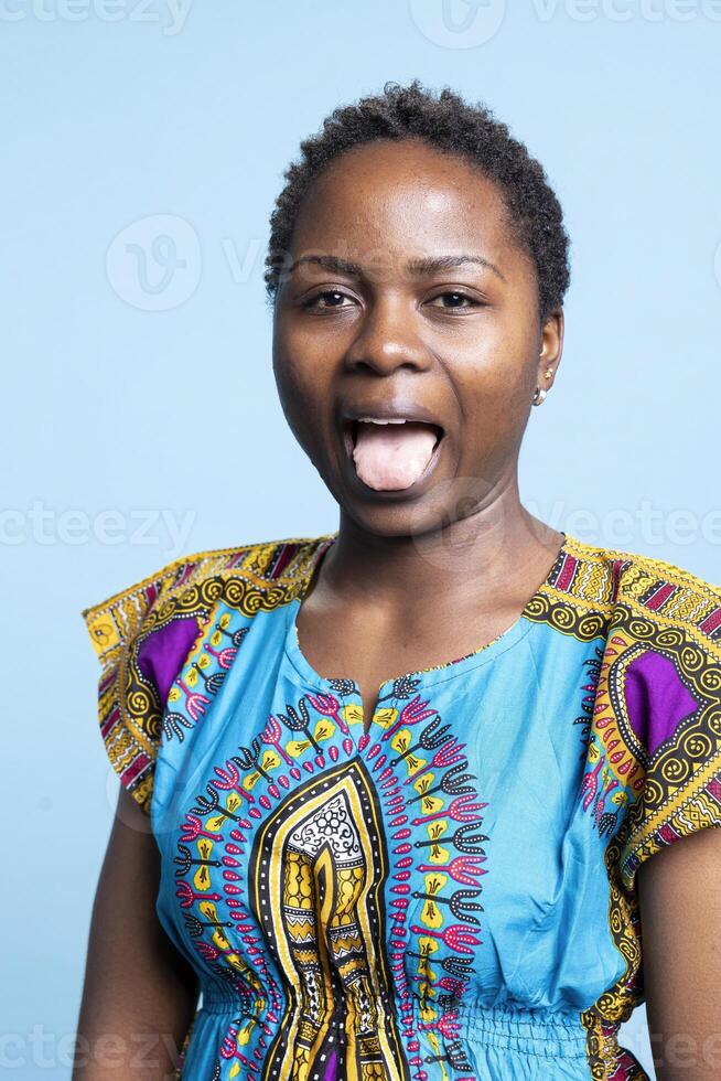 African american joyful person sticking tongue out on camera, fooling around and having fun. Young woman feeling confident and optimistic, making silly goofy facial expressions in studio. photo