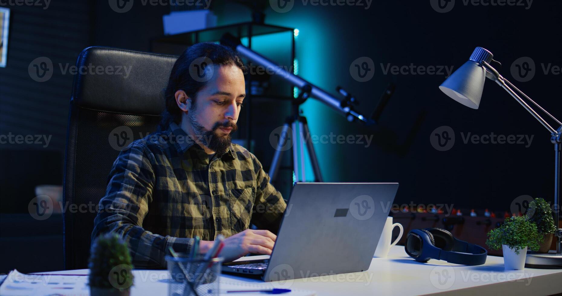 Smiling man sitting at home office desk and typing on laptop keyboard, solving tasks. Happy remote worker using notebook to send emails to business partners from living room, camera B photo