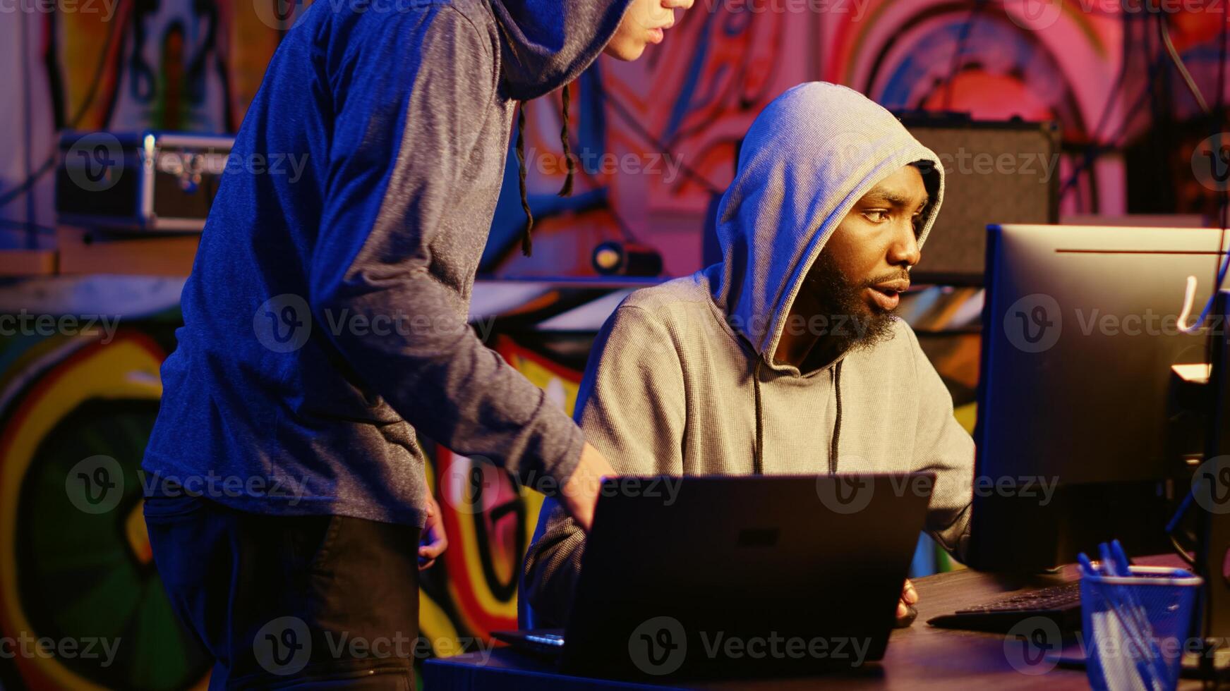Hacking group new member receiving guidance from knowledgeable rogue programmer acting as his mentor in ghetto abandoned hideaway. Master hacker teaches script kiddie how to deploy viruses photo