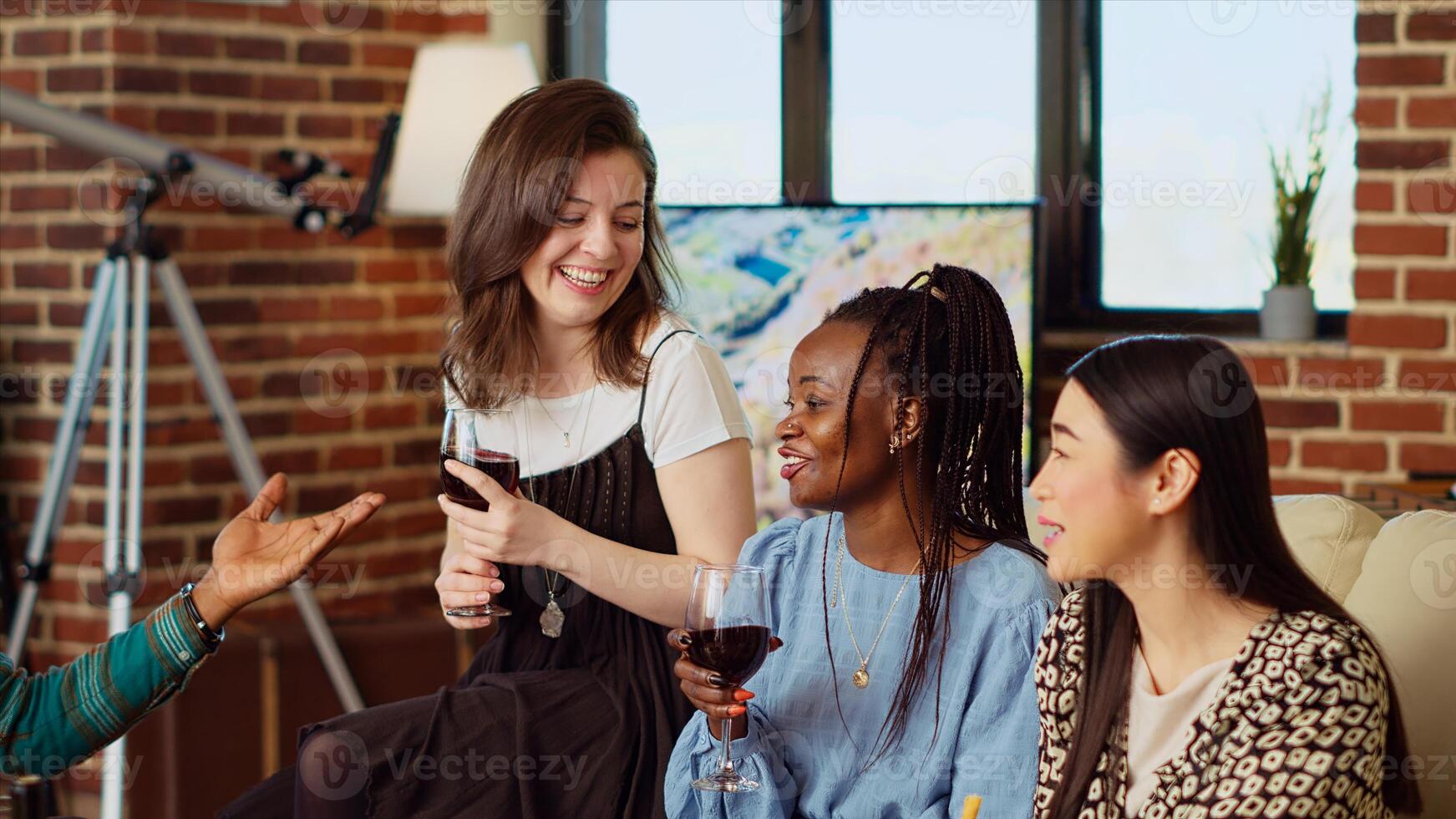 Best friends laughing together, enjoying alcohol beverages and gourmet cheese platter in cozy apartment living room. BIPOC group of people having lively conversation at home, entertaining each other photo