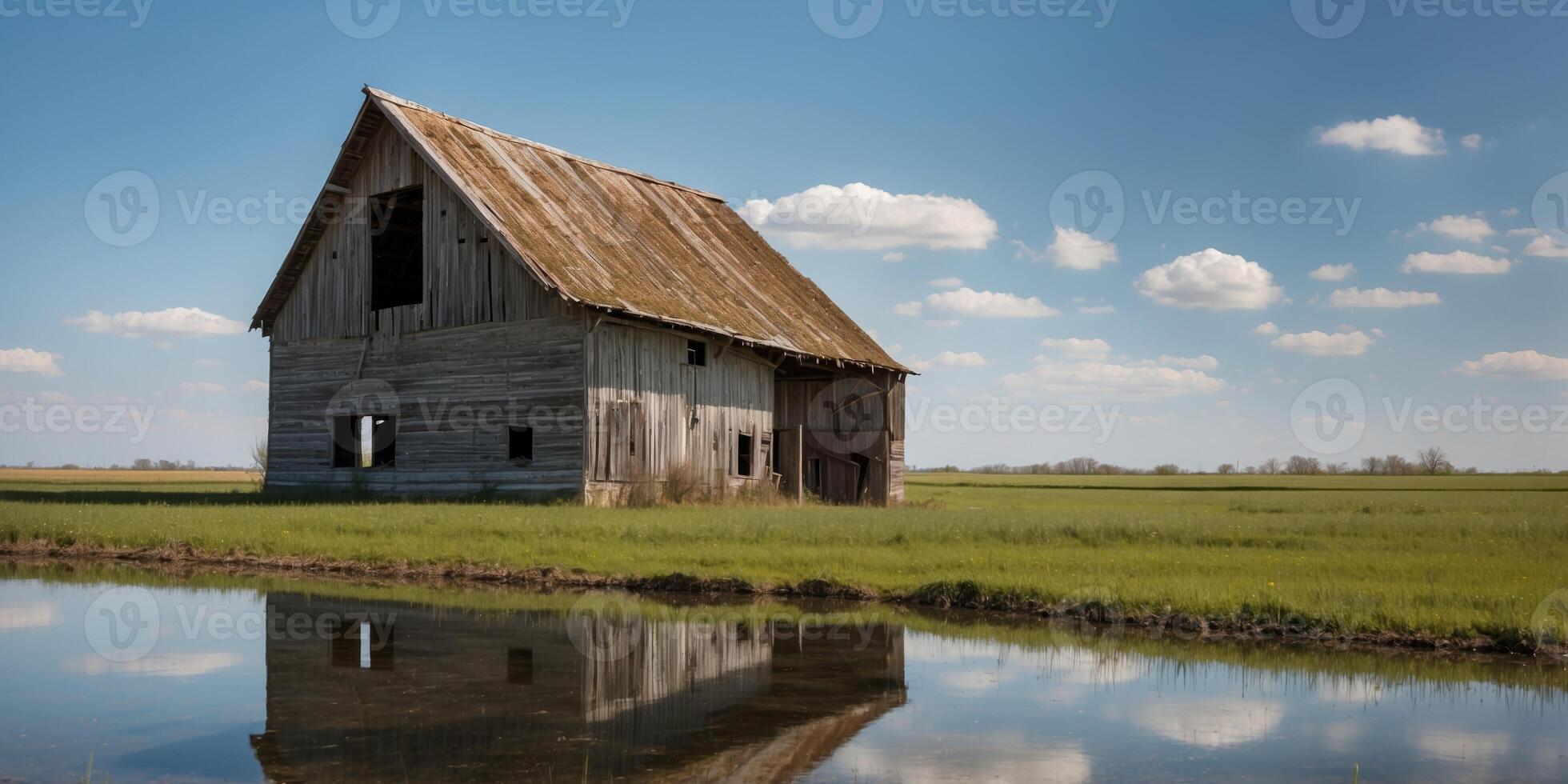 Weathered barn with a deteriorating roof in a green field near a blue water ditch on a sunny spring day. photo