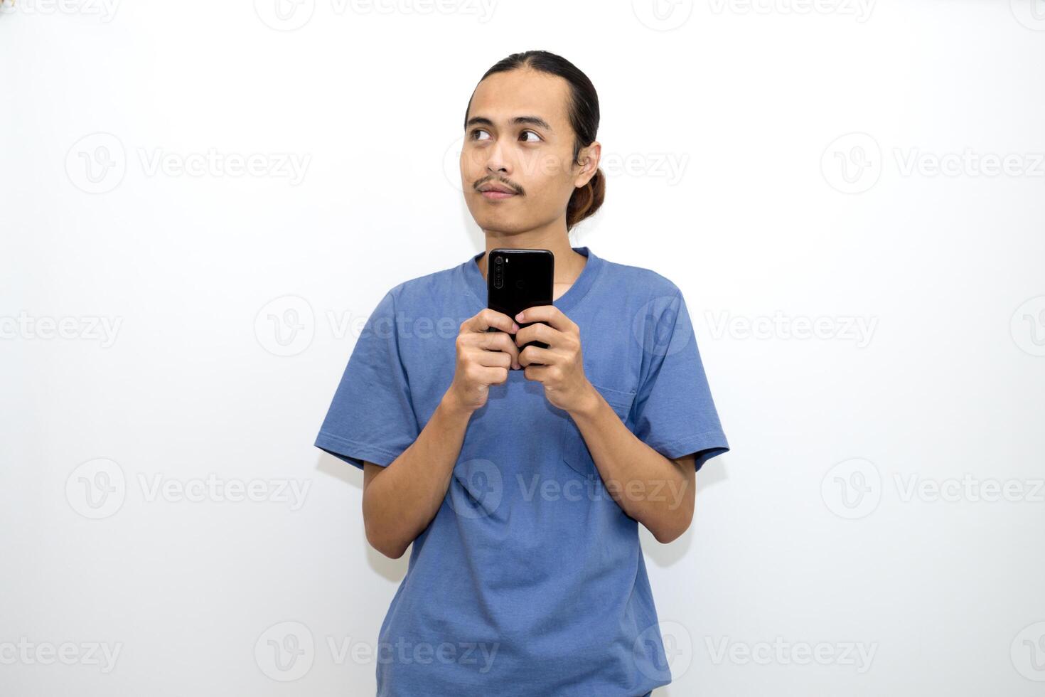 Adult Asian man wearing dark blue t-shirt showing thinking gesture, and holding mobile phone. isolated on white background photo