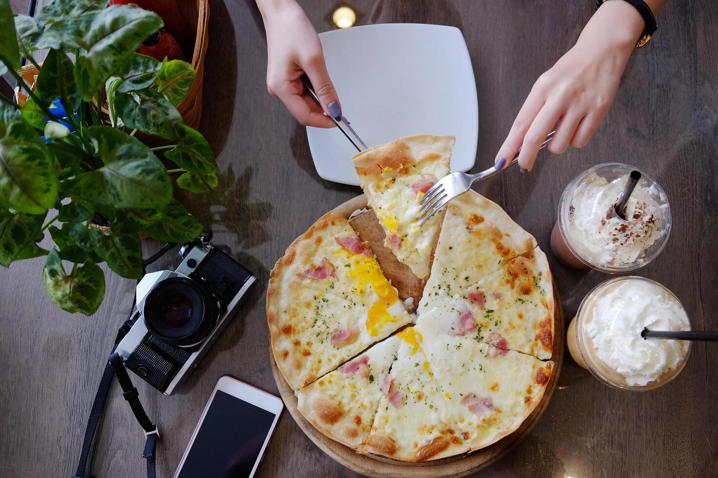 Top view for woman with knife and fork taking piece of delicious ham cheese pizza with vintage film camera and smartphone on wooden table in cafe and restaurant photo