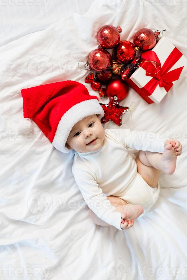 Christmas toddler in Santa hat sitting on bed with gift box and festive baubles photo