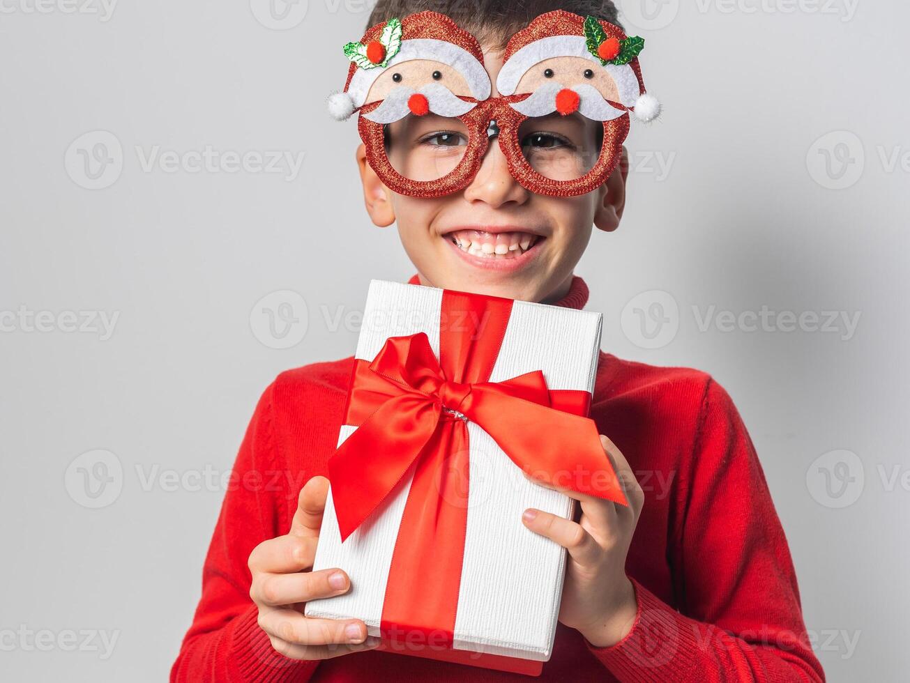 Close-up Smiling little boy wearing funny glasses in form of Christmas Santa Claus holding Xmas gift while standing in a white background, dressed in warm red xmas sweater photo