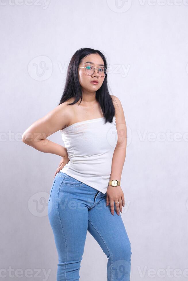 sexy young woman indonesia beautiful wearing glasses sharp to the camera hands holding waist and thighs wearing white tube top jeans standing isolated white background. for content beauty, fashion photo