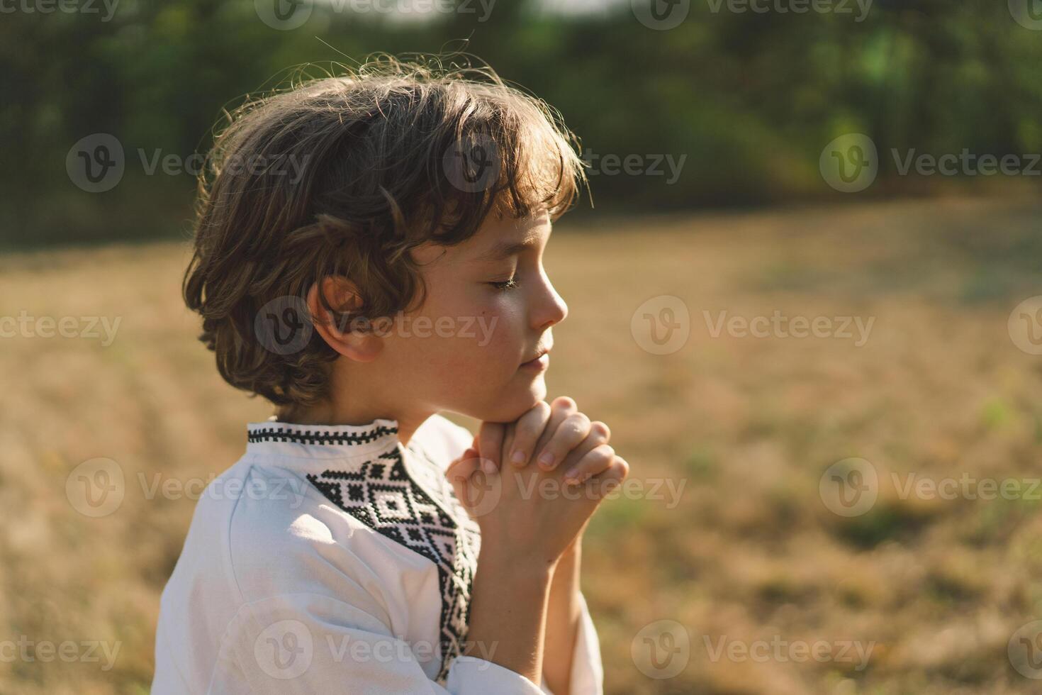 A young child wearing a traditional embroidered shirt quietly prays outdoors photo