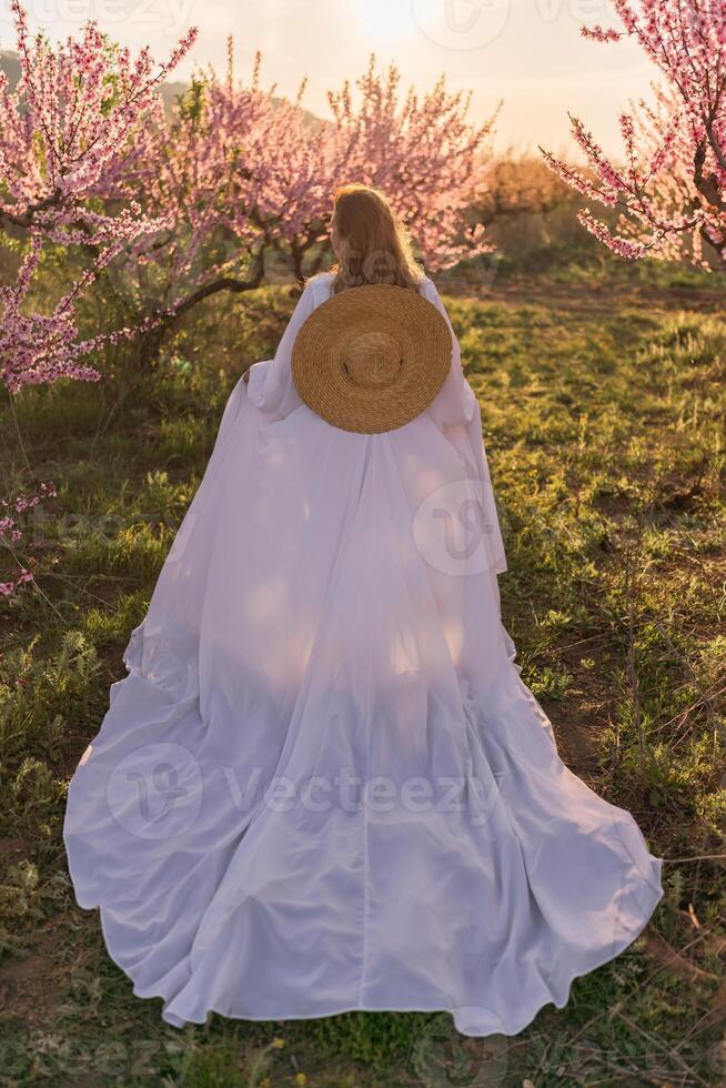 Woman blooming peach orchard. Against the backdrop of a picturesque peach orchard, a woman in a long white dress and hat enjoys a peaceful walk in the park, surrounded by the beauty of nature. photo