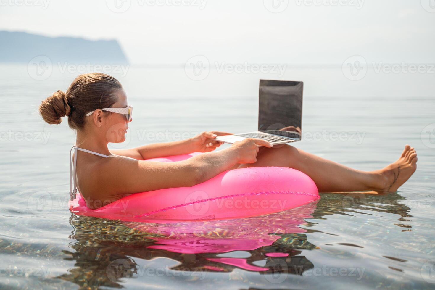 A woman is sitting in a pink inflatable raft on a lake, using a laptop. Concept of relaxation and leisure, as the woman is enjoying her time outdoors while working on her laptop. photo
