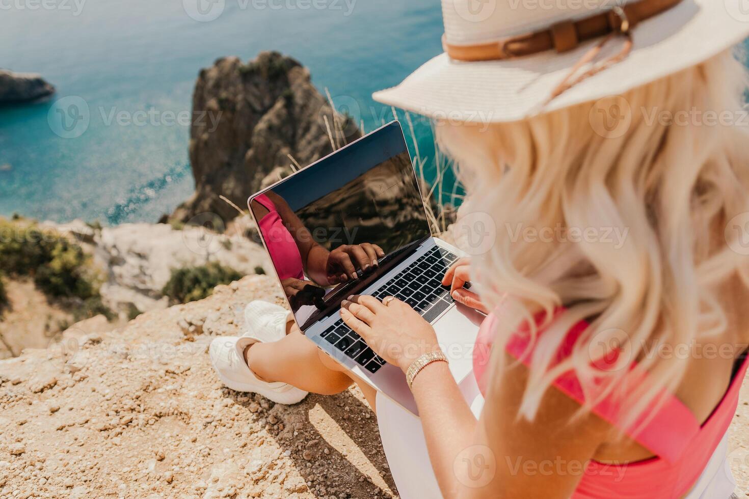 A woman is sitting on a rock by the ocean, typing on her laptop. Concept of relaxation and leisure, as the woman enjoys the beautiful view of the water while working on her computer. photo