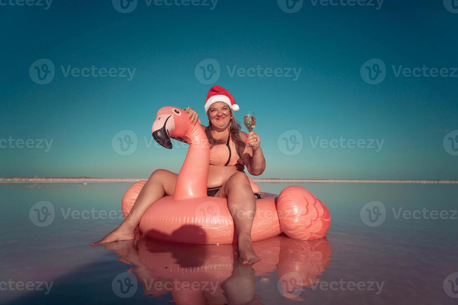 Santa Flamingo Christmas woman Riding Inflatable Flamingo Pink Lake. A woman wearing a Santa hat sits on a pink flamingo float in a body of water. He is smiling. photo