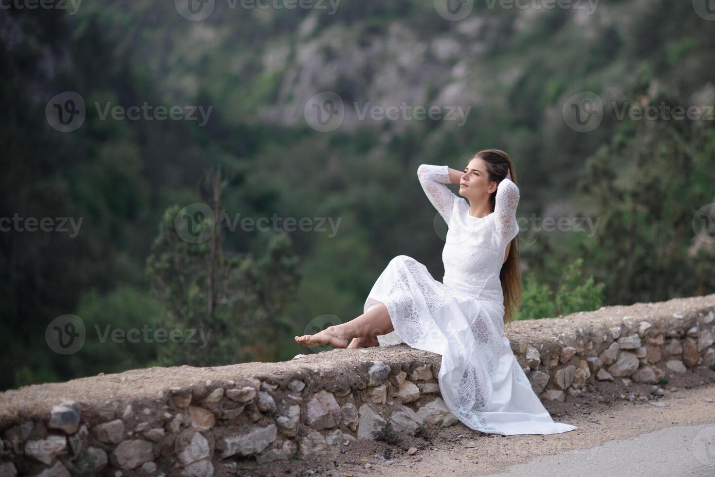 A woman in a white dress is sitting on a rock by a road. She is looking up at the sky. photo
