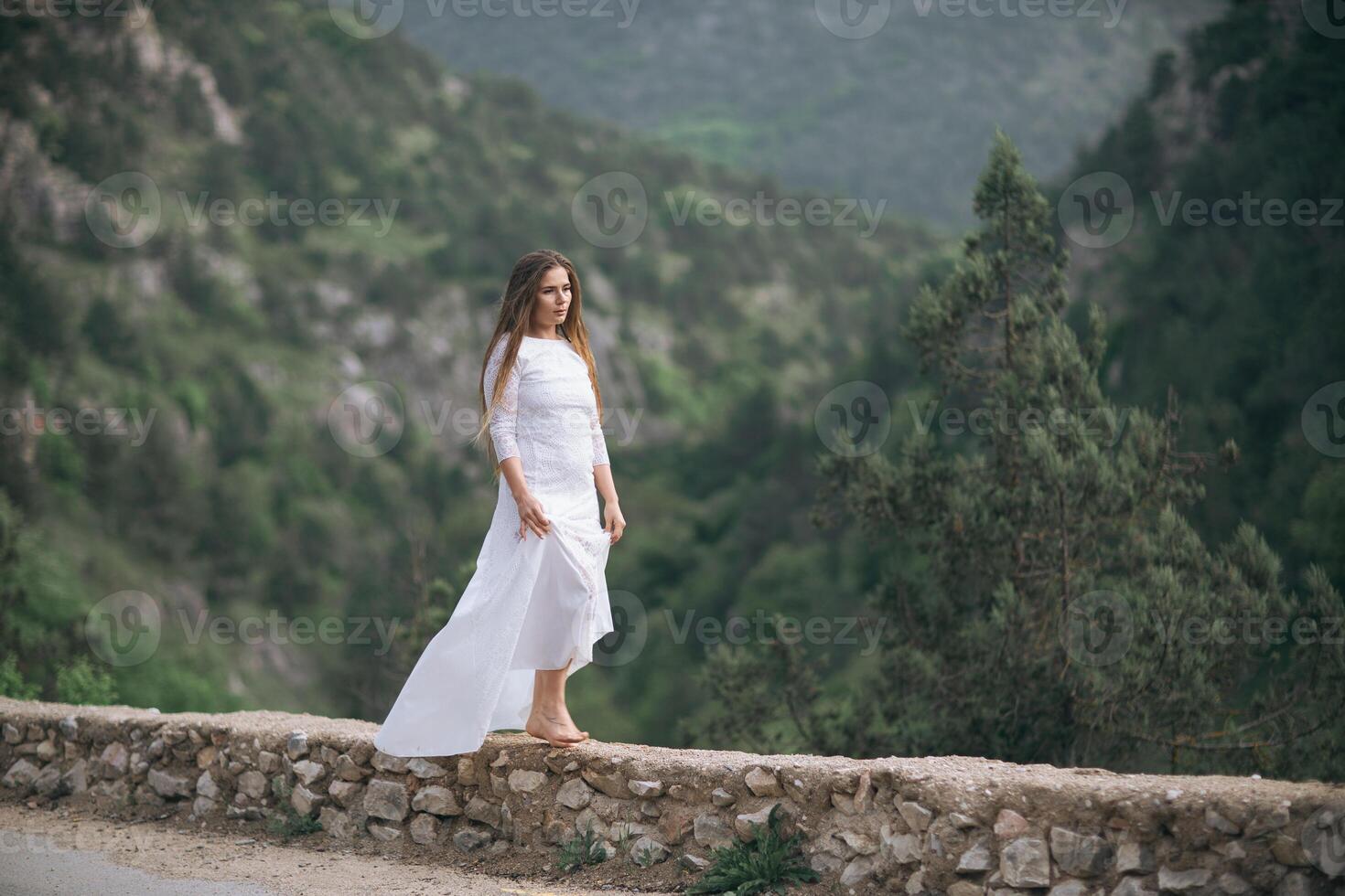 A woman in a white dress is walking on a stone wall. The image has a serene and peaceful mood, as the woman is alone and surrounded by nature. photo