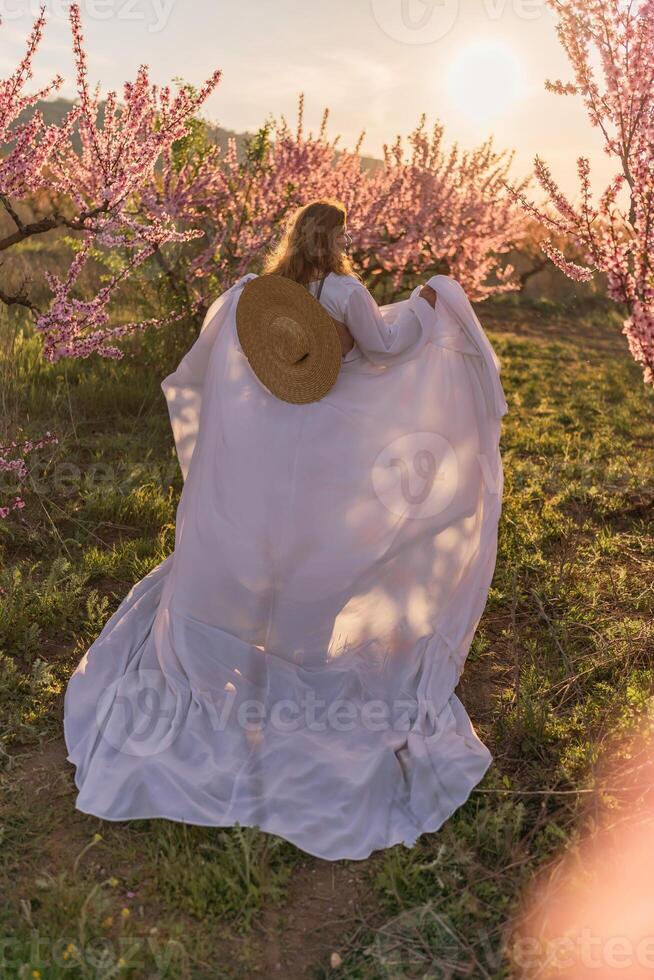 Woman blooming peach orchard. Against the backdrop of a picturesque peach orchard, a woman in a long white dress and hat enjoys a peaceful walk in the park, surrounded by the beauty of nature. photo