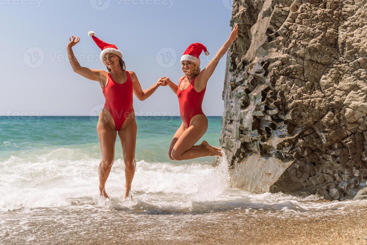 Women Santa hats ocean play. Seaside, beach daytime, enjoying beach fun. Two women in red swimsuits and Santa hats are enjoying themselves in the ocean waves and raising their hands up. photo