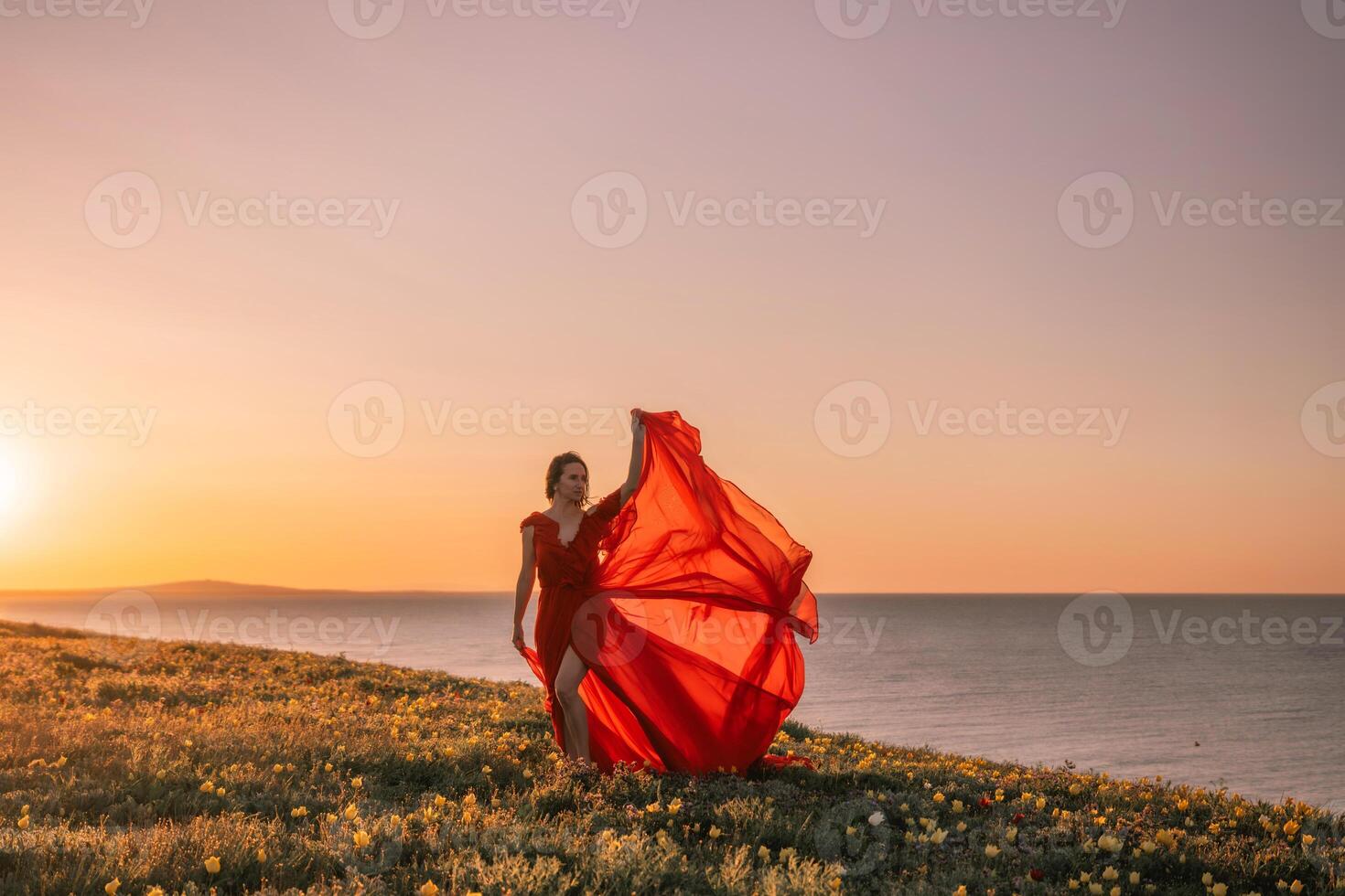 A woman in a red dress is standing on a grassy hill overlooking the ocean. The sky is a beautiful mix of orange and pink hues, creating a serene and romantic atmosphere. photo