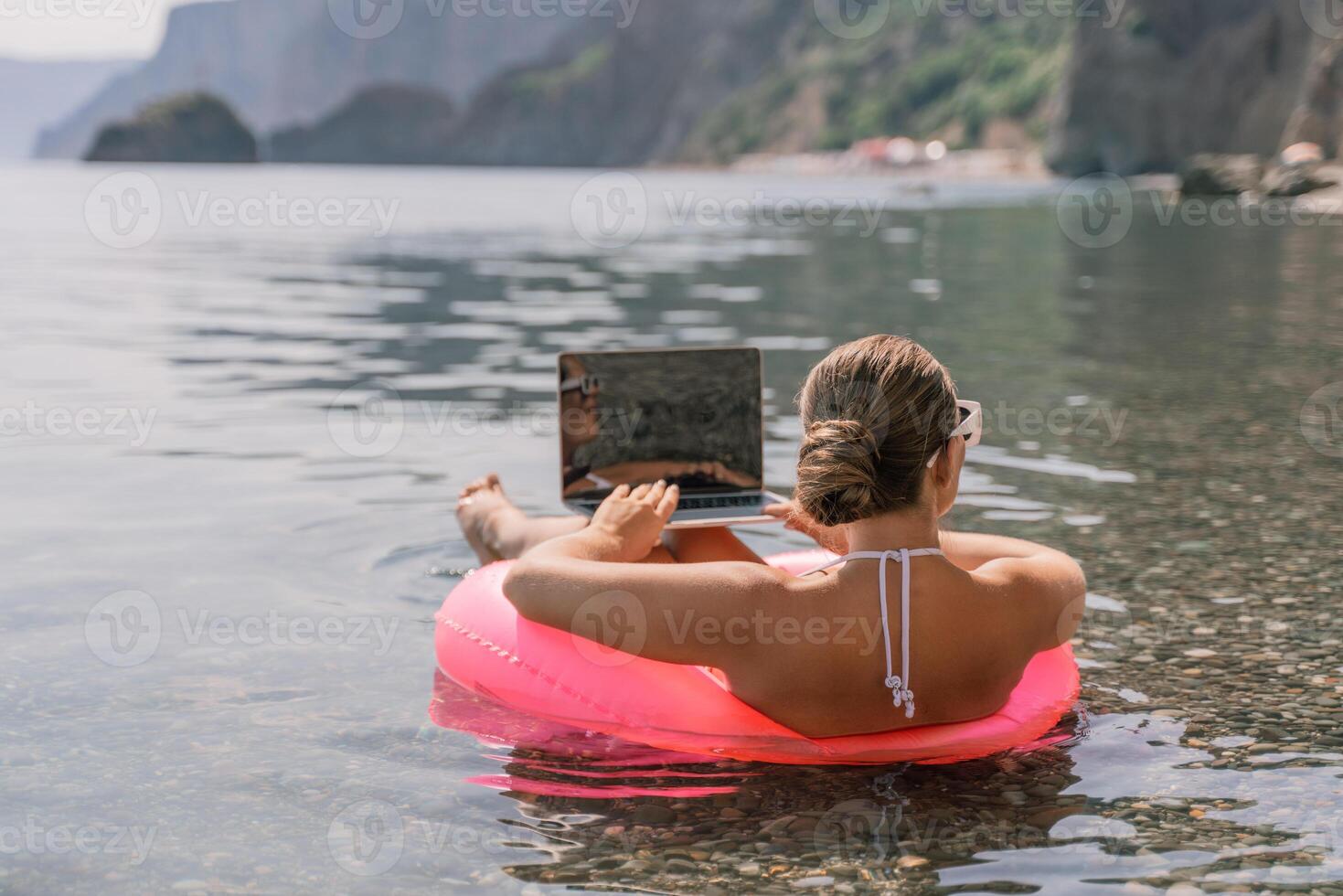 A woman is sitting on a pink inflatable raft in the water, using a laptop. Concept of relaxation and leisure, as the woman is enjoying her time in the water while working on her laptop. photo