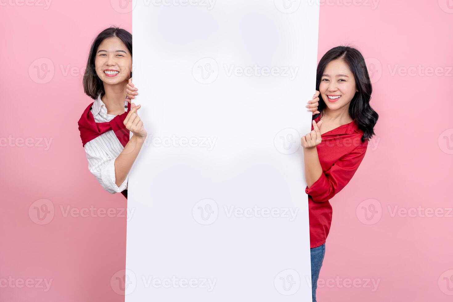 Two beautiful Asian girls standing behind the white blank banner or empty copy space advertisement board and showing mini heart sign photo