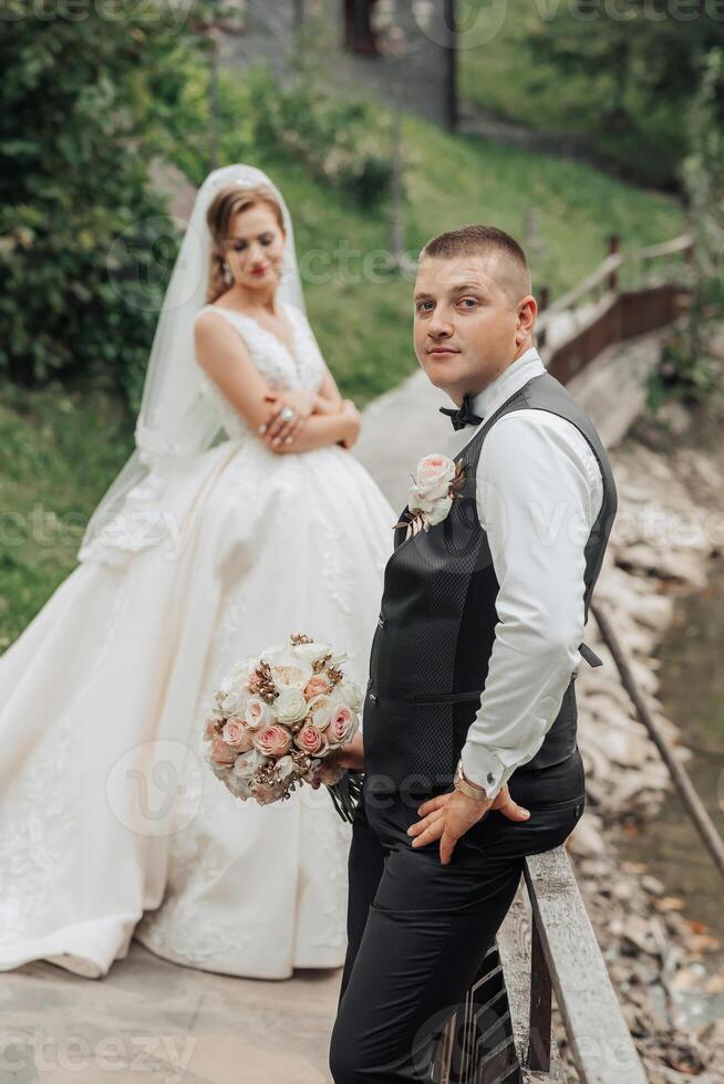 A bride and groom pose for a picture on a bridge. The bride is wearing a white dress and the groom is wearing a black vest. Scene is happy and celebratory, as the couple is getting married photo
